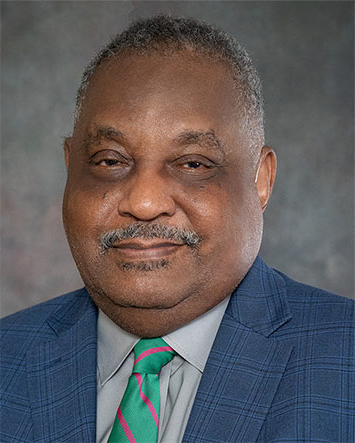 Mr. 詹姆斯·肯德里克, an African American male with salt and pepper hair and a goatee, grins at the camera wearing a blue plaid suit with a grey collared shirt and a mint and fushcia striped tie.