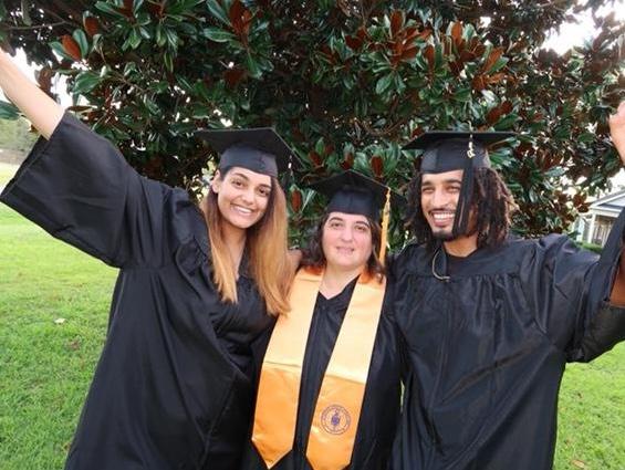 Smiling African American male with dreadlocks wearing a black graduation gown, 金色流苏的黑色毕业帽, gold necklace and a blacks shirt; hand is spread out; center: Caucasian female with brown hair wearing a black graduation gown, black graduation cap with a gold tassel and a gold stole with a logo, 左:微笑, female with golden brown hair wearing a black graduation cap, black graduation gown; hand is spread out; group is standing in front of a tree