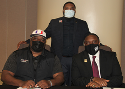 Three African American males wearing face masks and business attire posing for a photo; two males are sitting, 一只雄性站着, 坐着的两位男性都把手放在前面的桌子上.