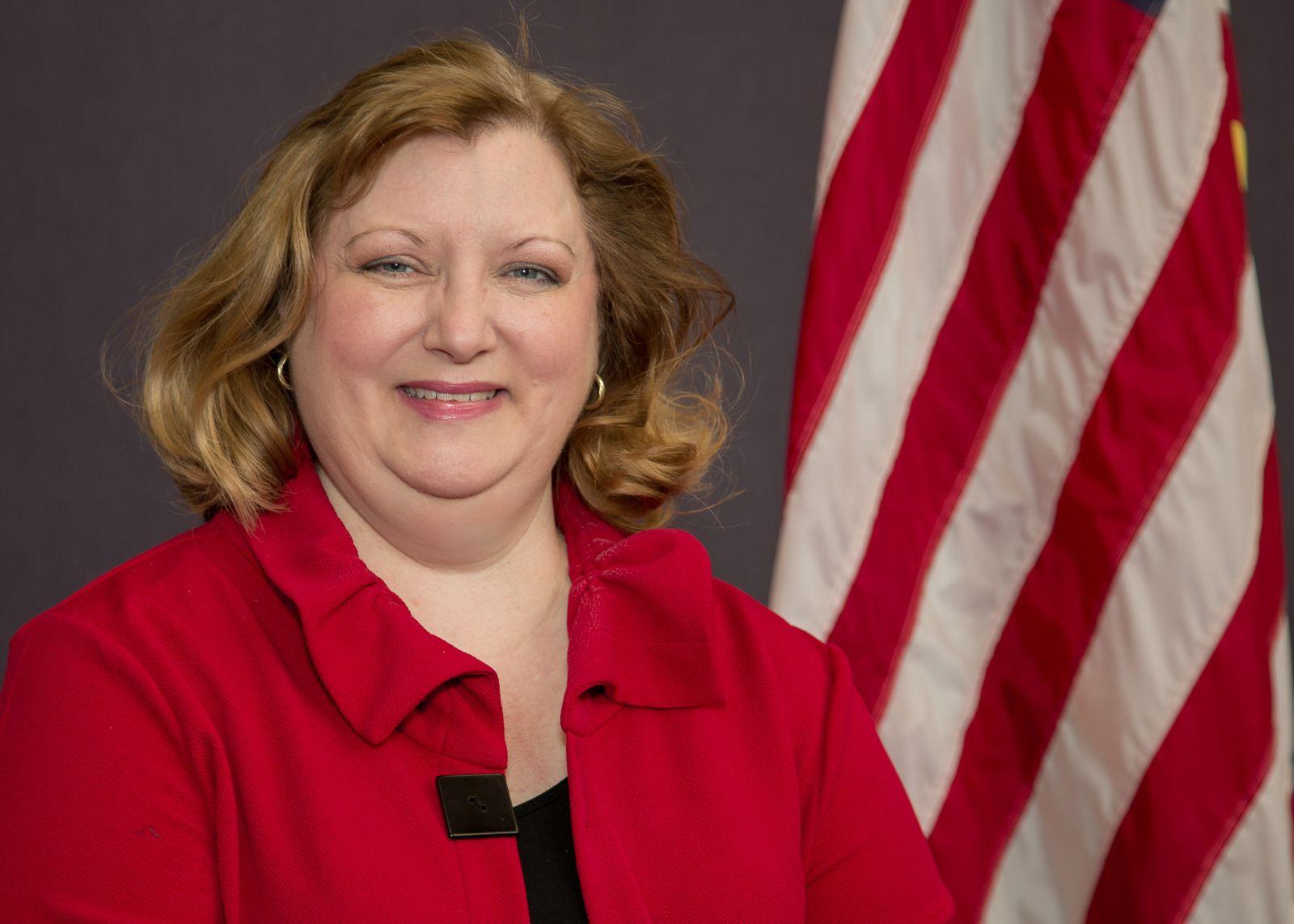 一个金发女郎, 白色的白种人, 颊酸式焦磷酸钠, is wearing a bright red collared jacket over a black shirt and is pictured with the United States flag standing in the back left against a grey background.