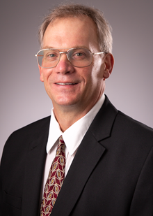 Caucasian male, smiling, wearing glasses, whit collared shirt, black suit jacket, red tie with design