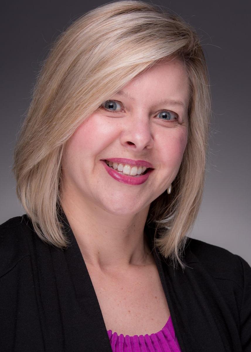 Smiling Caucasian female wearing a black blazer and purple shirt posing for a photo against a dark gray background